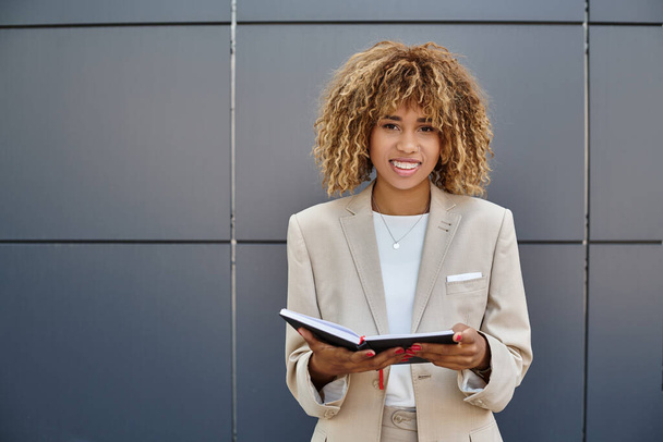 pozytywny afrykański amerykański bizneswoman w formalnym ubraniu trzymając jej notatnik w pobliżu biurowca - Zdjęcie, obraz