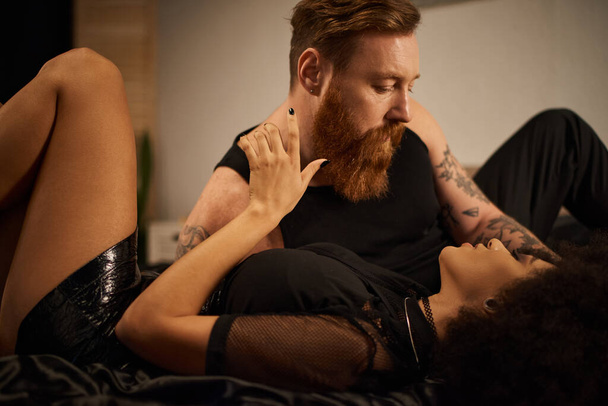 бородатий чоловік з татуюваннями спокушає дівчину в спальні, палкий момент мультикультурної пари - Фото, зображення