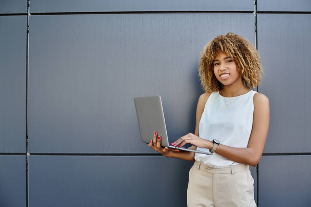 glücklich lockige afrikanisch-amerikanische Freiberuflerin steht mit ihrem Laptop in der Nähe der grauen Wand eines Bürogebäudes - Foto, Bild