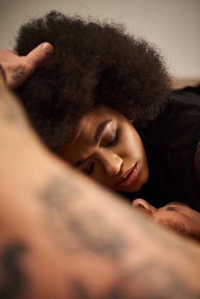 Αφροαμερικανή γυναίκα με σγουρά μαλλιά ξαπλωμένη δίπλα στο αγόρι της με τατουάζ στο θολή χέρι - Φωτογραφία, εικόνα