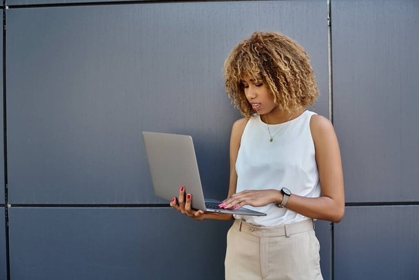 mujer de negocios afroamericana rizada con frenos de pie con su computadora portátil cerca de la pared gris del edificio - Foto, imagen