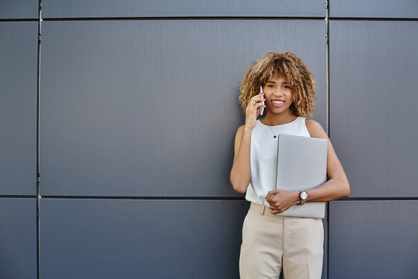 Ευτυχισμένη νεαρή επαγγελματίας, αφροαμερικανή γυναίκα με φορητό υπολογιστή κάνει μια κλήση σε γκρι φόντο - Φωτογραφία, εικόνα