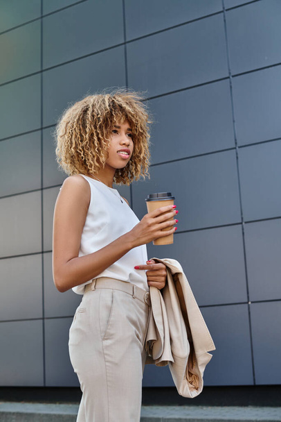 Stylish and curly african american businesswoman with coffee standing against a modern building - Photo, Image