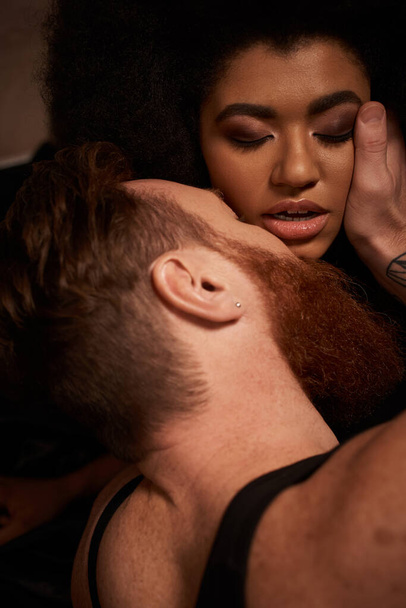 hombre barbudo con tatuajes tocando la cara de la mujer americana africana rizada mientras la seduce en la cama - Foto, Imagen