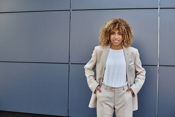 młody szczęśliwy czarny bizneswoman w garniturze stojąc z rękami w kieszeniach przeciw szary budynek - Zdjęcie, obraz