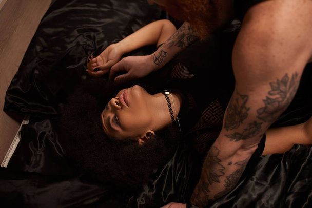 ベッドの上のカーリー髪の若いアフリカ系アメリカ人女性を誘惑するタトゥー付きのひげ付き男 - 写真・画像