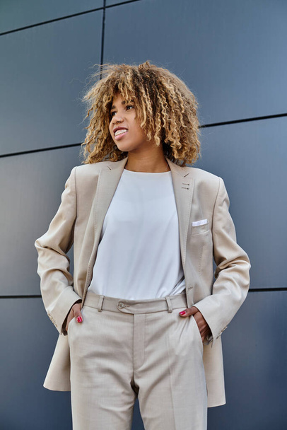 feliz mulher de negócios afro-americana de terno de pé com as mãos nos bolsos contra edifício cinza - Foto, Imagem