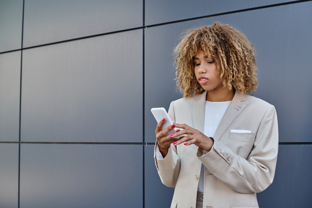 joven mujer de negocios afroamericana con el pelo rizado de pie en traje y utilizando su teléfono inteligente - Foto, Imagen