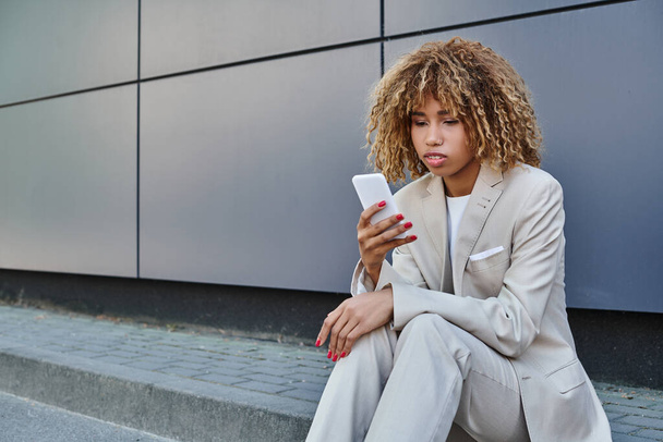 young african american businesswoman with curly hair standing in suit and using smartphone outside - Zdjęcie, obraz