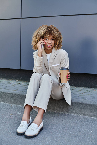 Αφροαμερικανή γυναίκα με κοστούμι κάθεται σε στηθαίο με καφέ για να πάει και να μιλήσει στο smartphone της - Φωτογραφία, εικόνα