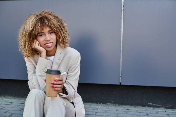 happy african american woman in suit sitting on parapet with coffee to go and looking at camera - Photo, Image