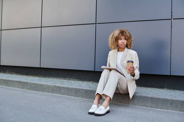 Elegante professionista, donna afro-americana con caffè e cartella seduta vicino alla parete dell'ufficio - Foto, immagini