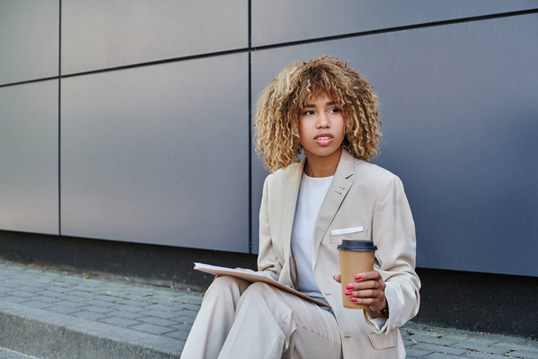 Elegante profesional, mujer afroamericana con café y carpeta sentado en la pared gris de la oficina - Foto, imagen