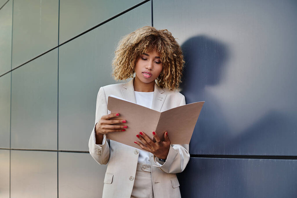focalisé afro-américaine bouclé femme d'affaires examen de la paperasserie dans le dossier près de mur de bureau gris - Photo, image