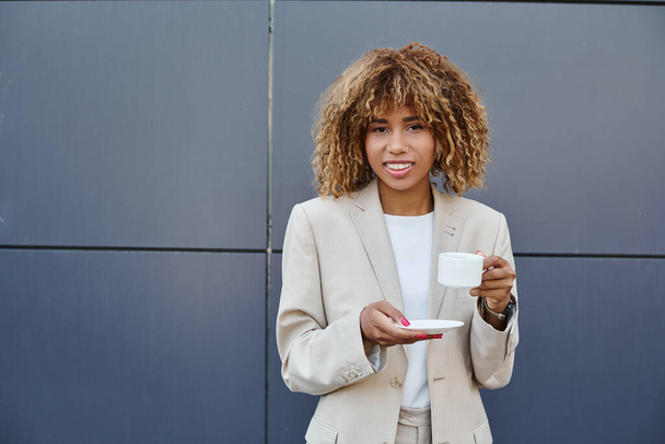cheerful african american businesswoman standing with cup of coffee near entrance of office building - Zdjęcie, obraz