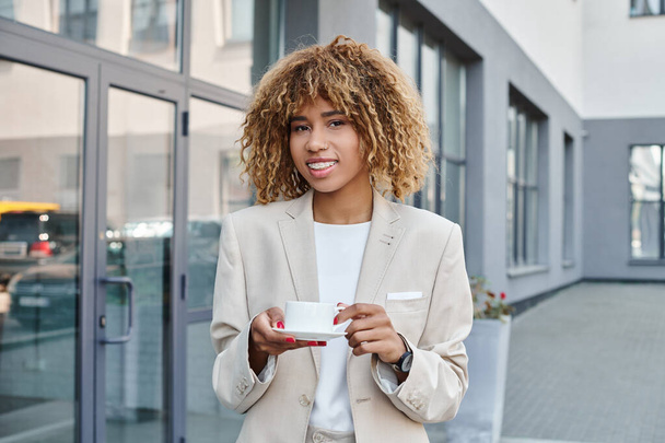 lachende Afro-Amerikaanse zakenvrouw in de 20, staande met een kop koffie in de buurt van kantoorgebouw - Foto, afbeelding