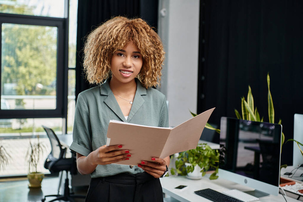 felice donna d'affari afroamericana sulla ventina, in piedi con il caffè per andare e la cartella in ufficio - Foto, immagini