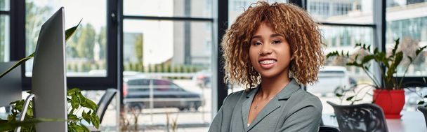 щаслива афроамериканська бізнес-леді, що сидить за столом перед монітором комп'ютера в офісі, банер - Фото, зображення