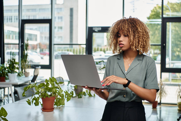 Ασχολείται με την εργασία νεαρή Αφροαμερικανή επιχειρηματίας χρησιμοποιώντας φορητό υπολογιστή, ενώ στέκεται σε καλά φωτισμένο γραφείο - Φωτογραφία, εικόνα