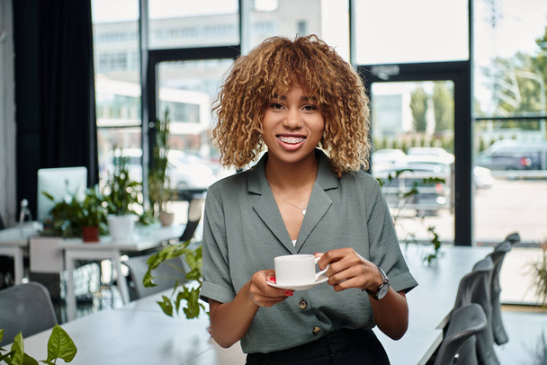 happy african american businesswoman smiling and enjoying coffee break in contemporary office - Photo, Image