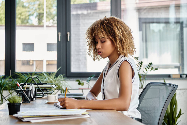 молодая африканская американская бизнесвумен с вьющимися волосами сидит за рабочим столом и делает заметки - Фото, изображение