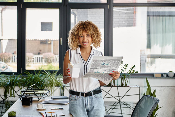 happy african american woman holding asian takeaway meal in carton box and newspaper in office - Photo, Image