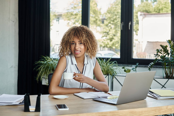 happy african american businesswoman with braces holding cup of coffee beside laptop in office - Photo, Image