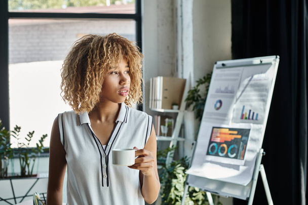 curly african american businesswoman with braces holding cup of coffee beside charts and graphs - Photo, Image