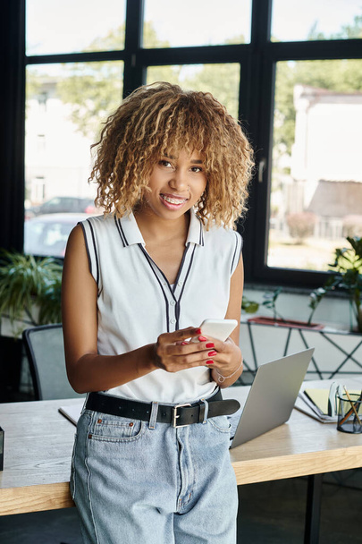 curly and happy african american businesswoman with smartphone staying connected in sunlit office - Photo, Image