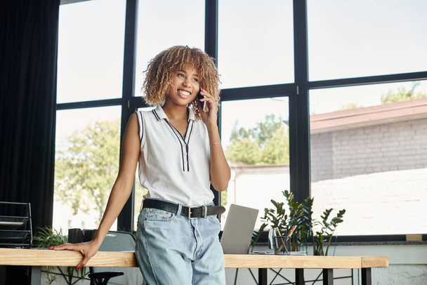 rizado y feliz afroamericana mujer de negocios teniendo llamada telefónica en la oficina moderna iluminada por el sol - Foto, imagen