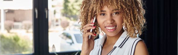 rizado y alegre mujer de negocios afroamericana que tiene llamada telefónica en la oficina moderna soleada, pancarta - Foto, Imagen