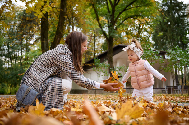 Anne ve tatlı bebek, sonbahar parkında yerde yapraklarla yürüyorlar. Annem parkta küçük bir çocukla vakit geçiriyor. # İyi bir anne-çocuk ilişkisi dışında #. - Fotoğraf, Görsel