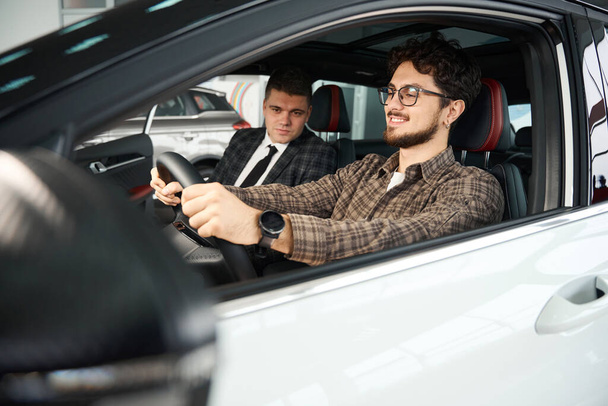 Улыбающийся человек выбирает новую машину с профессиональным продавцом, сидящим в автомобильном обсуждении моделей автомобилей - Фото, изображение