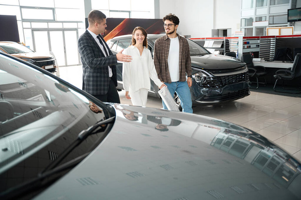 Веселая пара, посещающая автосалон или выставку, беседующая с продавцом о покупке нового автомобиля - Фото, изображение
