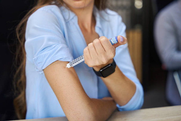 Selective focus of blurred cropped sick businesswoman taking injection with syringe in hand during work in coworking office. Concept of workaholism and working burnout - Photo, Image