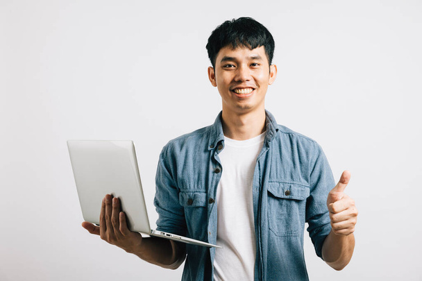 A successful young Asian man, holding a laptop, smiles with confidence, showing a thumbs-up for approval. Studio shot isolated on white background, a gesture of agreement. - Photo, Image