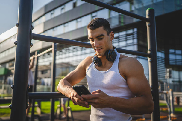 Ένας άνδρας καυκάσιος νεαρός άνδρας σταθεί στο υπαίθριο ανοιχτό γυμναστήριο του πάρκου κατάρτισης χρησιμοποιούν κινητό τηλέφωνο smartphone με ακουστικά στέλνουν μηνύματα γραπτών μηνυμάτων ή περιηγηθείτε στο διαδίκτυο online app για την κατάρτιση πραγματικό πρόσωπο αντιγραφή χώρου - Φωτογραφία, εικόνα