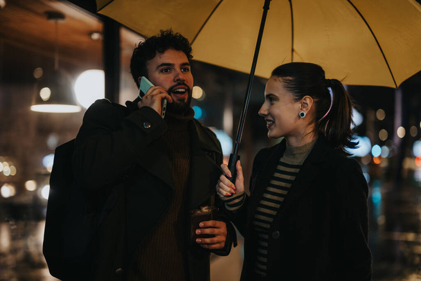 Elegante pareja que se enfrenta a un día lluvioso, usando un cajero automático para retirar dinero mientras sostiene un paraguas y mantiene una conversación. - Foto, imagen