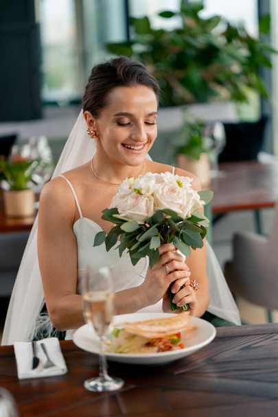 портрет красивой улыбающейся невесты на свадьбе в ресторане во время празднования любви с букетом цветов - Фото, изображение