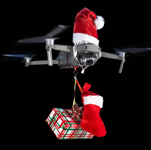 Koncepcja drona nosząca czapkę Mikołaja i trzymająca zapakowany prezent dla dostawy na czarnym tle. - Zdjęcie, obraz