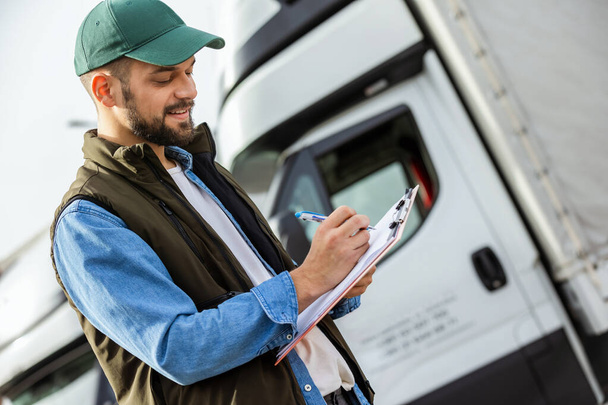 Motorista masculino confiante feliz em pé na frente em seu caminhão enquanto escreve algo em seu papel. - Foto, Imagem