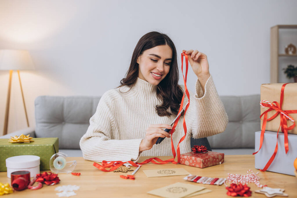 Vrolijke vrouw in gebreide trui verpakking kerstcadeaus met papier, schaar en kleurrijke linten. Feestelijke feestvoorbereidingen. - Foto, afbeelding