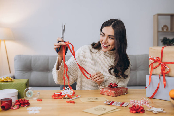 Boldog nő kötött pulóverben karácsonyi ajándékokat csomagolva papírral, ollóval és színes szalagokkal. Ünnepi előkészületek. - Fotó, kép