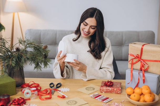Frau packt Weihnachtsgeschenkschachteln mit Emotionen ein oder aus, hautnah. Unvorbereitete Geschenke auf dem Tisch mit Dekorationselementen und Artikeln Weihnachten oder Neujahr. - Foto, Bild