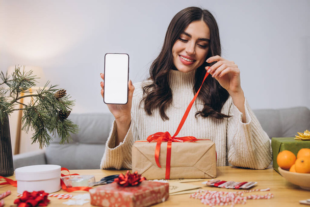 Hermosa mujer feliz haciendo envoltura de regalo de Año Nuevo o Navidad mientras sostiene el fondo del teléfono inteligente con pantalla en blanco con espacio para copiar o burlarse en casa - Foto, imagen