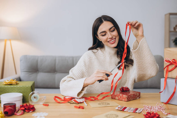 Glückliche Frau im Strickpullover, die Weihnachtsgeschenke mit Papier, Schere und bunten Bändern einwickelt. Festliche Feiertagsvorbereitungen. - Foto, Bild