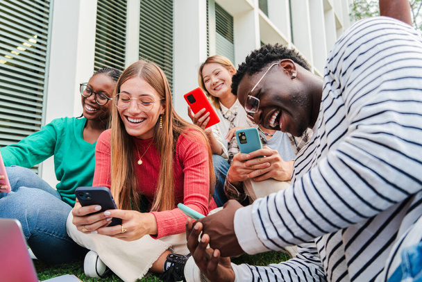 Группа улыбающихся молодых мультиэтнических старшеклассников, использующих мобильные телефоны. Парни и женщины смотрят социальные сети со своими смартфонами, наслаждаясь вместе, сидя на лужайке университетского городка - Фото, изображение