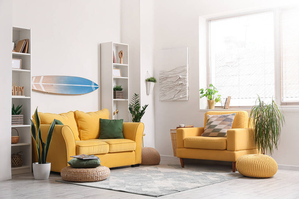 Interno di soggiorno luminoso con tavola da surf, divano e poltrona gialli - Foto, immagini
