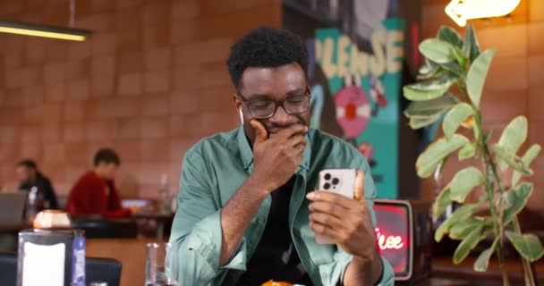 Bell'uomo afroamericano con gli occhiali che indossa gli auricolari mentre parla online con qualcuno sul suo cellulare. Giovane ragazzo si diverte durante la conversazione a distanza. Dispositivo tecnologico. - Filmati, video