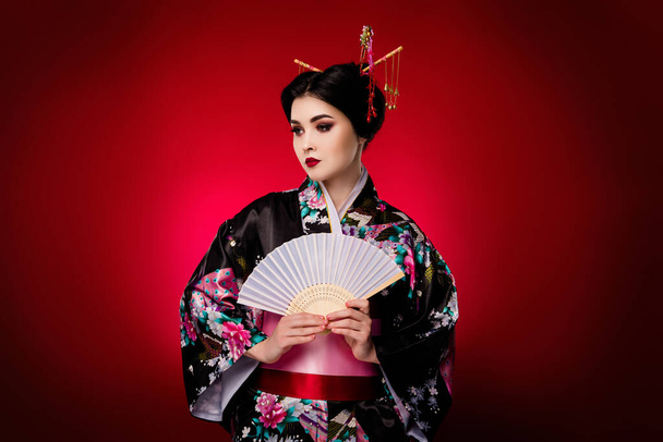 Foto de glamour asiático dama anime teatro personaje vestido kimono celebrar ventilador aislado en rojo degradado color fondo. - Foto, imagen